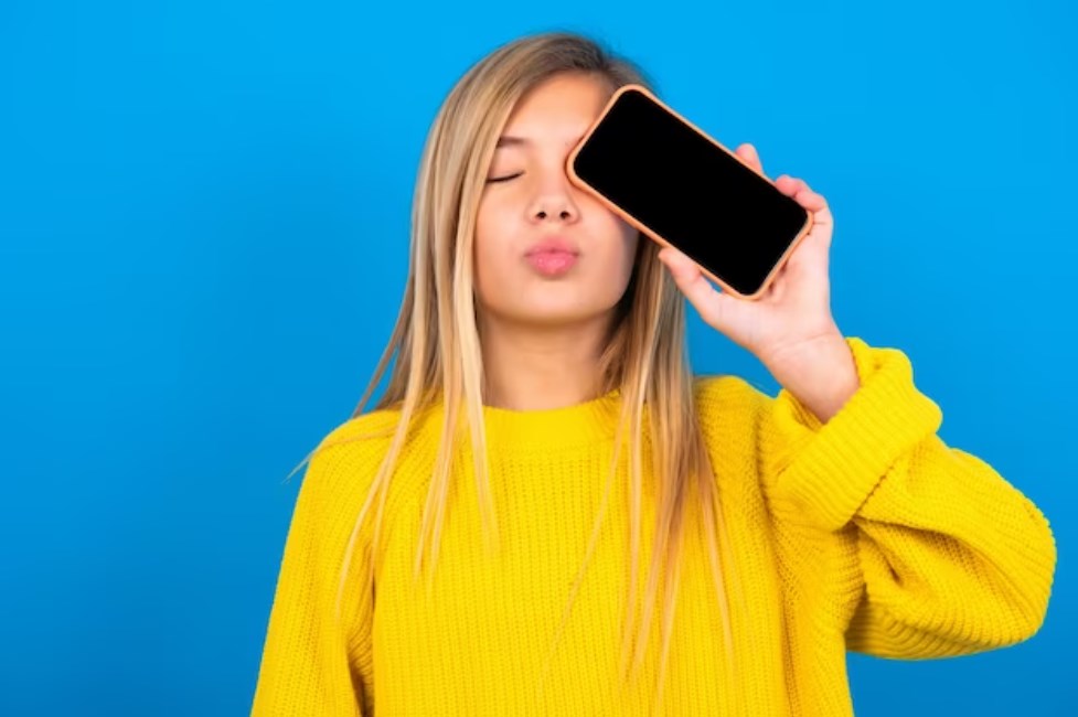 a teen girl wearing a yellow sweater and holding a smartphone сovering her left eye with her lips pouted
